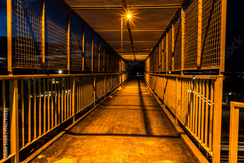 Night view of a pedestrian bridge with yellow lighting. Dramatic urban scene, suitable for city infrastructure, safety, or design themes. Striking contrast