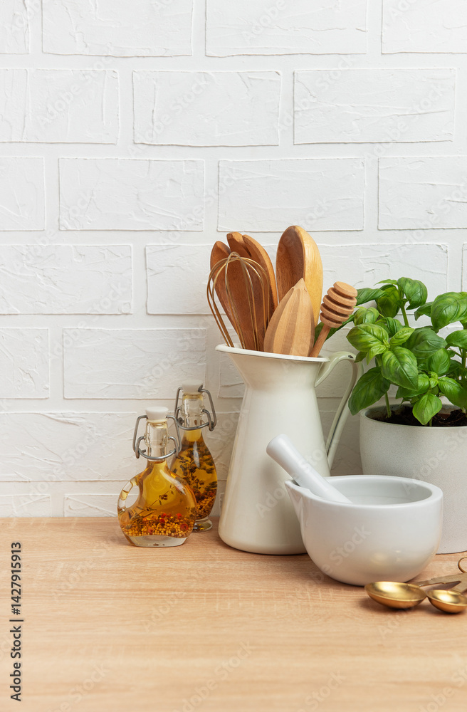Kitchen utensils, basil plant and oil bottles resting on wooden countertop in front of white brick wall