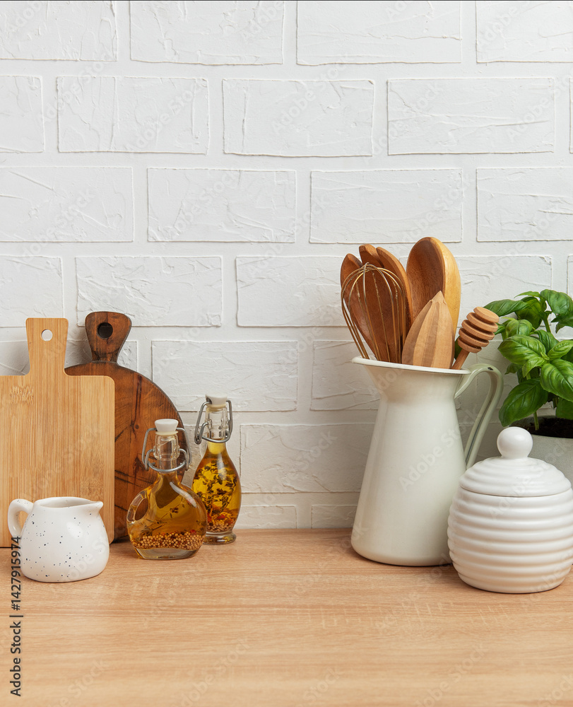 Kitchen utensils, basil plant and condiments resting on wooden countertop in front of white brick wall