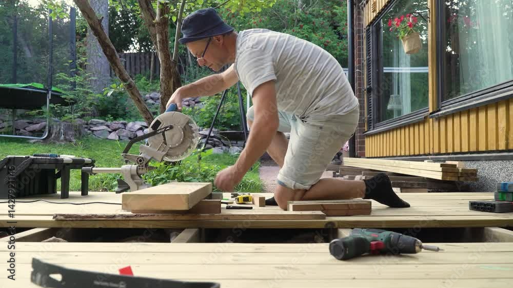 Man working on wooden deck construction outdoors using a circular saw, building a patio in backyard on a sunny day.