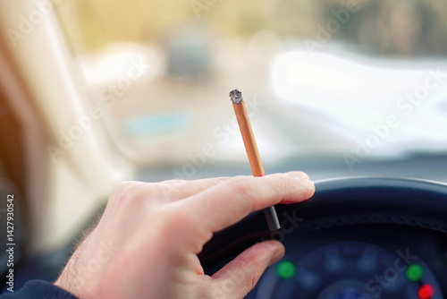 Close up of driver gripping the wheel with one hand while holding burning cigarette, unsafe driving habits, legal concerns, and potential traffic penalties. Smoking while driving, selective focus.