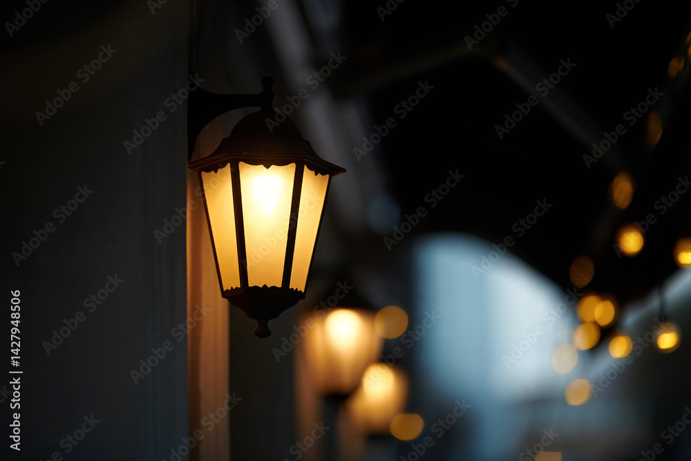 Glowing antique style lantern mounted on wall, providing warm illumination and an inviting ambiance in nighttime city setting with string lights in background. Nighttime atmosphere. Selective focus