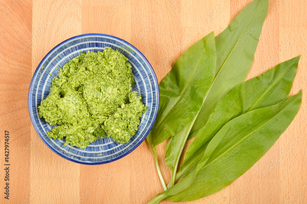 A blue and white bowl of vibrant green wild garlic (Allium ursinum) butter sits beside fresh wild garlic leaves on a wooden surface, captured from above