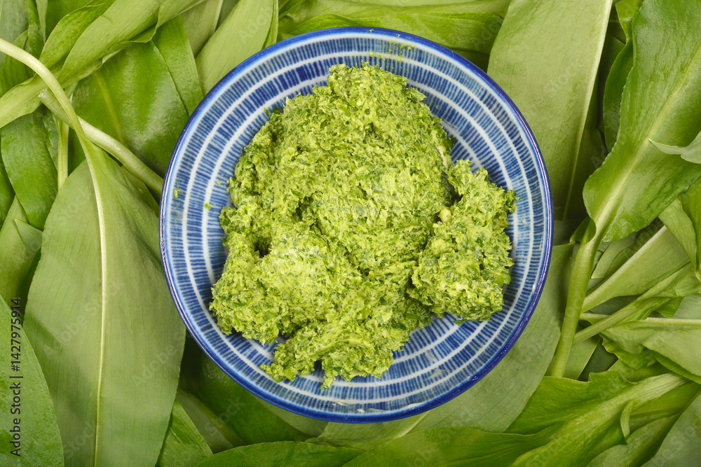A blue and white bowl filled with vibrant green wild garlic (Allium ursinum) butter is nestled amongst fresh wild garlic leaves in a top-down composition
