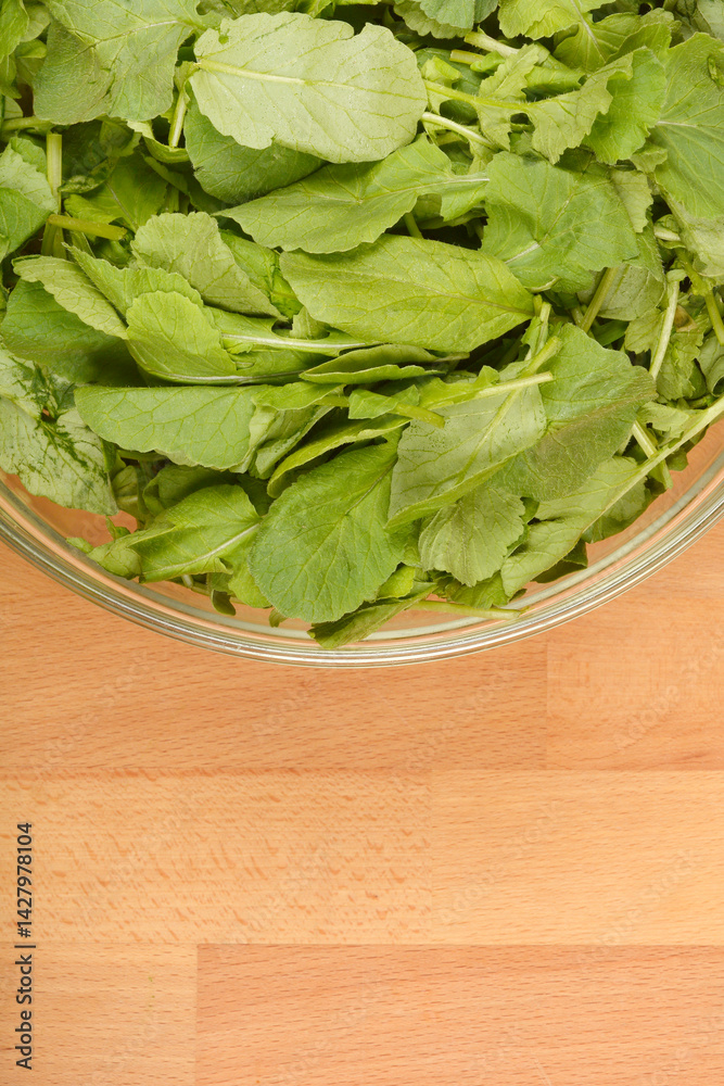 A clear glass bowl filled with fresh, vibrant green radish leaves sits on a light wooden surface, captured from a top-down perspective