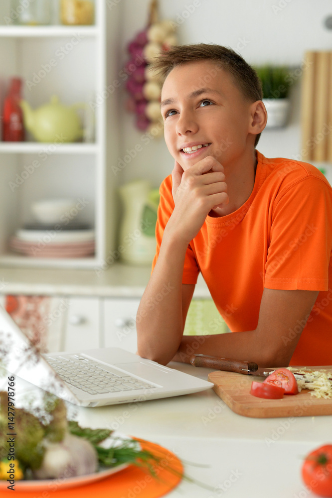 Teen boy thinking while working on laptop in kitchen