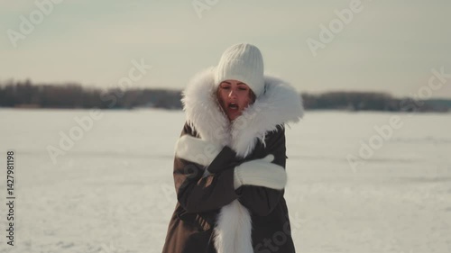 Freezing winter landscape with shivering woman wearing warm white hat, bundled against arctic cold, standing on snow-covered frozen lake shoreline