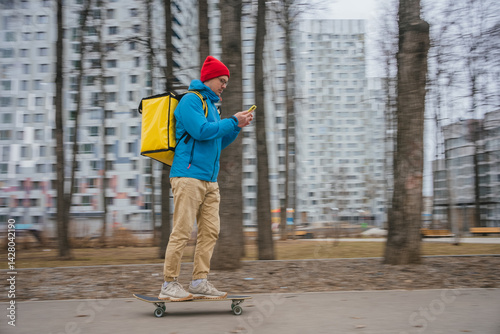 Young male food delivery courier with yellow thermal backpack riding skateboard and checking smartphone while delivering order through modern urban area on a cold day.