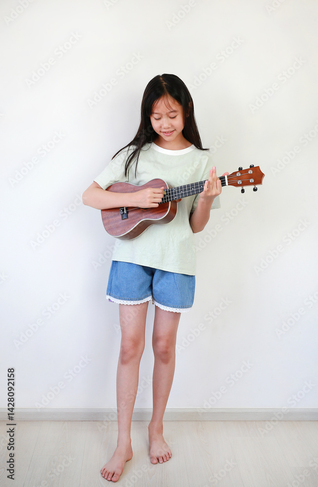 Asian girl child playing ukulele while standing in the white room. Full body photo.