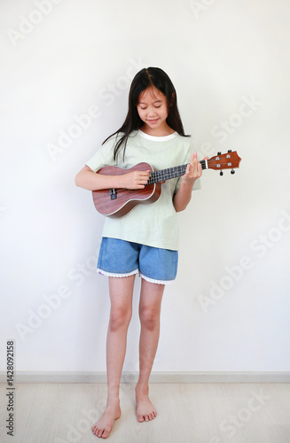 Obraz na płótnie Asian girl child playing ukulele while standing in the white room. Full body photo. w salonie