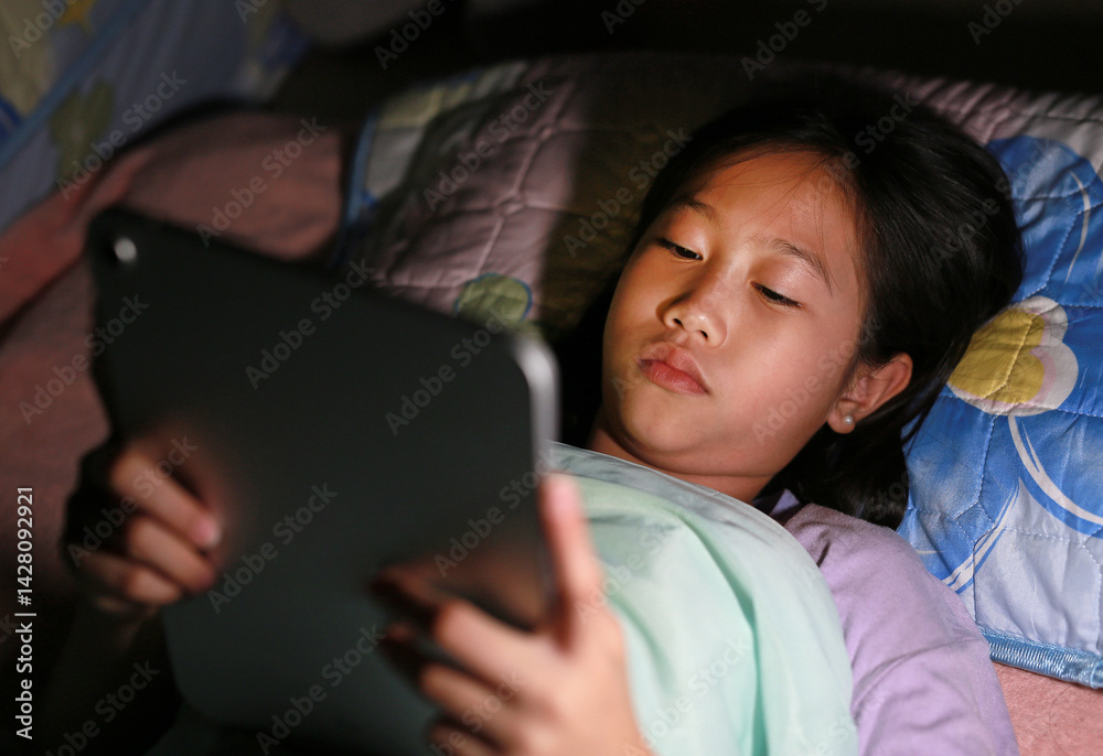 Portrait of Asian girl preteen playing digital tablet lying on bed at night.