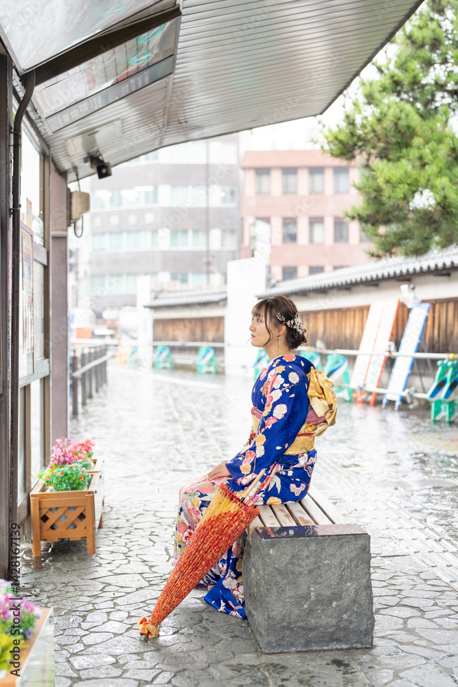 It's a rainy summer day. bus stop. A Japanese woman in a teenager wearing a blue kimono folded and placed it by her side, sitting on a bench.