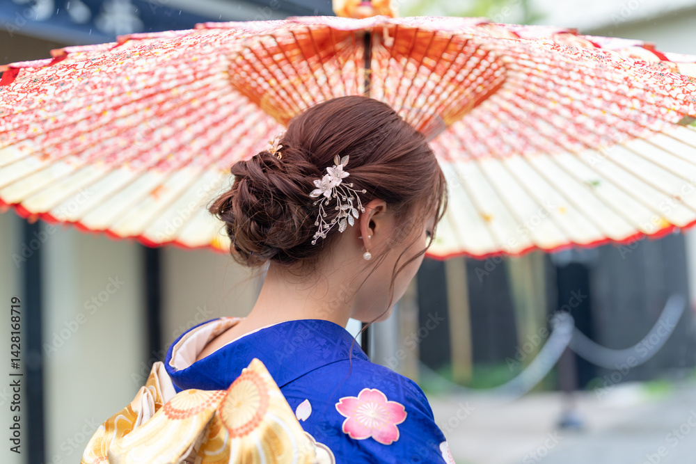 A local city in Japan on a rainy summer day. A Japanese woman wearing a blue teenage kimono holding a Japanese umbrella and climbing a slope. The back view.