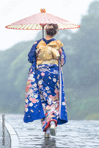 A local city in Japan on a rainy summer day. A Japanese woman wearing a blue teenage kimono holding a Japanese umbrella and climbing a slope. The back view.
