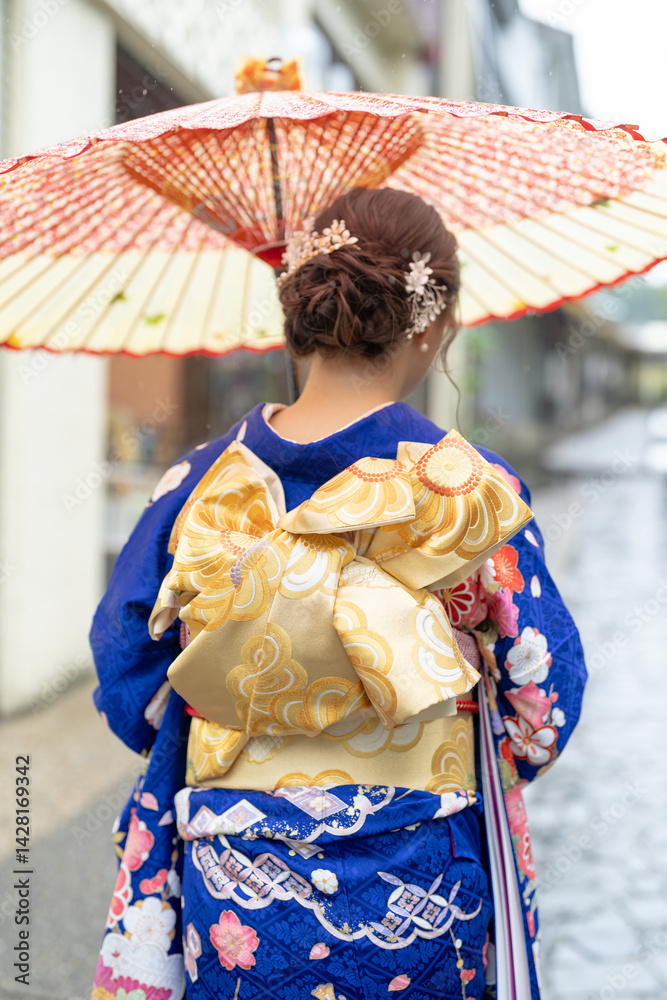 A local city in Japan on a rainy summer day. A Japanese woman wearing a blue teenage kimono holding a Japanese umbrella and climbing a slope. The back view.