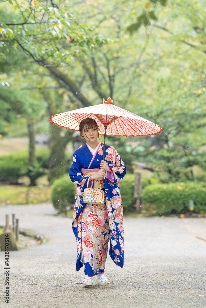 A local city in Japan on a rainy summer day. Japanese woman wearing a blue kimono for teenagers with a Japanese umbrella walking through a traditional garden.