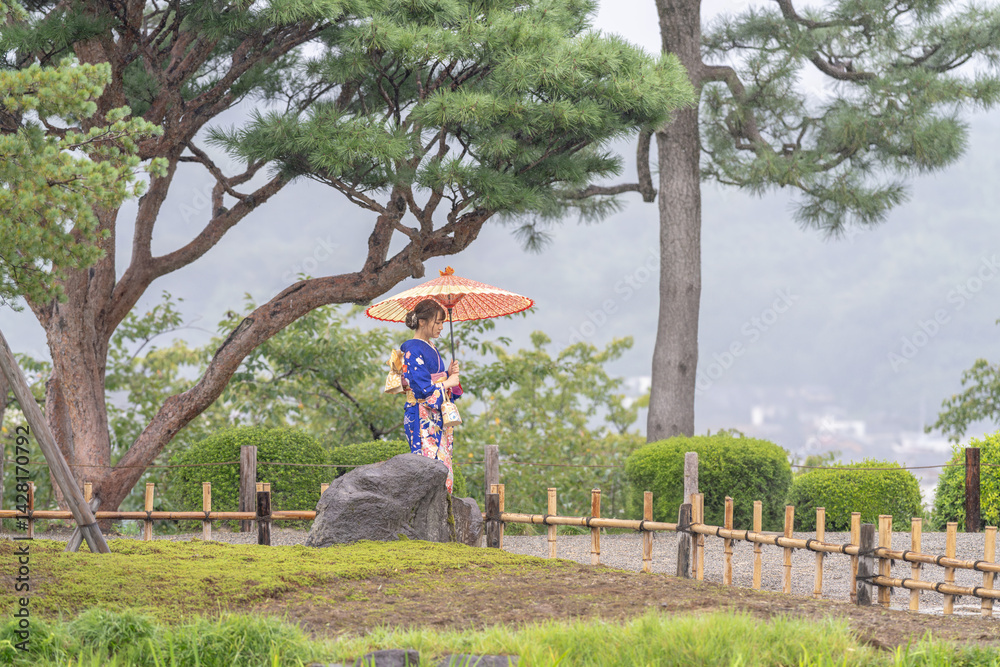 A local city in Japan on a rainy summer day. Japanese woman wearing a blue kimono for teenagers with a Japanese umbrella walking through a traditional garden.