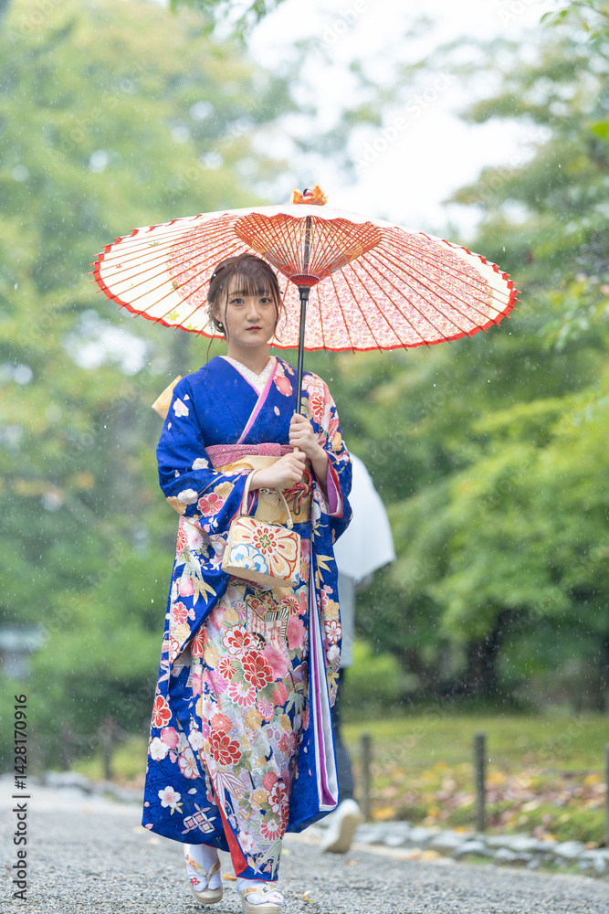 A local city in Japan on a rainy summer day. Japanese woman wearing a blue kimono for teenagers with a Japanese umbrella walking through a traditional garden.