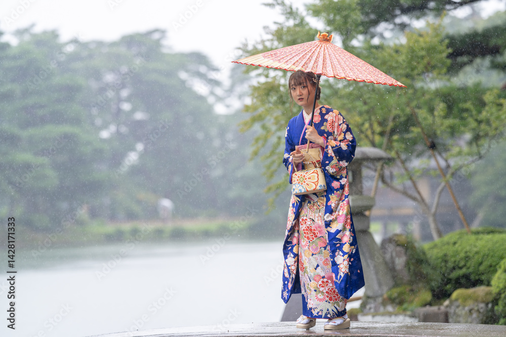 A local city in Japan on a rainy summer day. Japanese woman wearing a blue teenage kimono with a Japanese umbrella walking by a pond in a traditional garden.