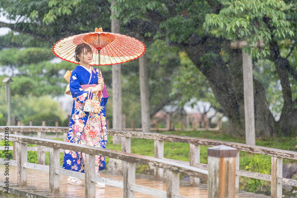 A local city in Japan on a rainy summer day. Japanese woman wearing a blue teenage kimono with a Japanese umbrella walking along a traditional garden bridge.
