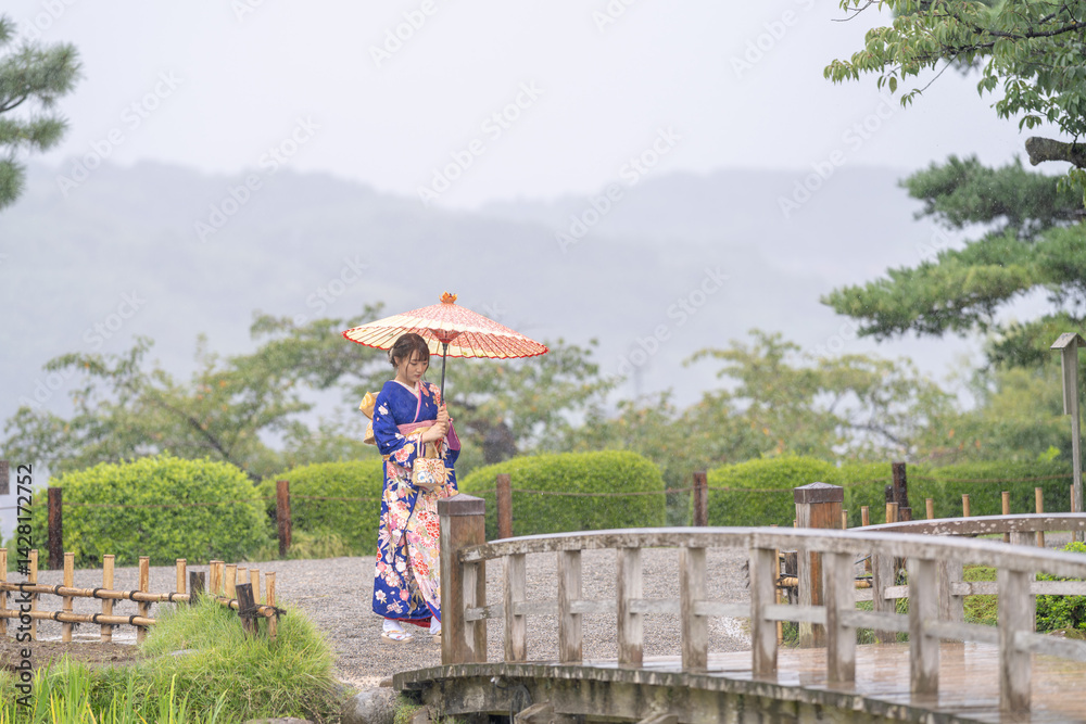 A local city in Japan on a rainy summer day. Japanese woman wearing a blue kimono for teenagers with a Japanese umbrella walking through a traditional garden.