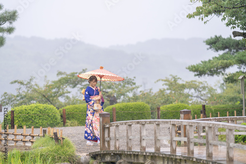 Obraz na płótnie A local city in Japan on a rainy summer day. Japanese woman wearing a blue kimono for teenagers with a Japanese umbrella walking through a traditional garden. w salonie