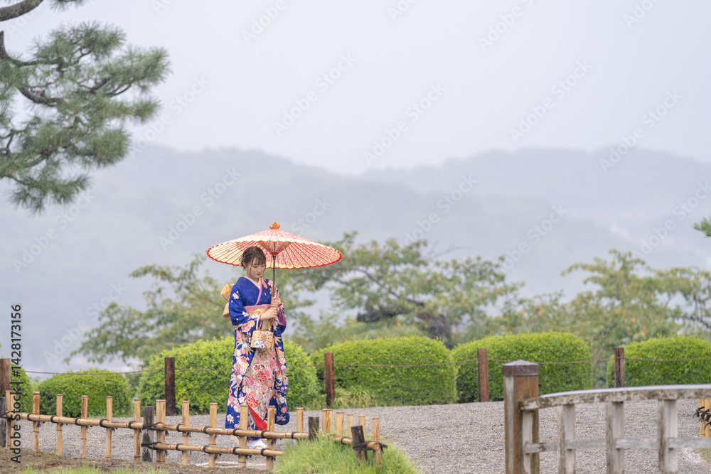A local city in Japan on a rainy summer day. Japanese woman wearing a blue kimono for teenagers with a Japanese umbrella walking through a traditional garden.