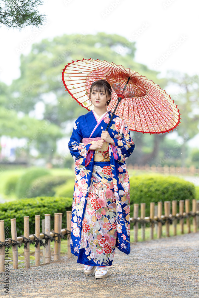 A local city in Japan on a rainy summer day. Japanese woman wearing a blue kimono for teenagers with a Japanese umbrella walking through a traditional garden.