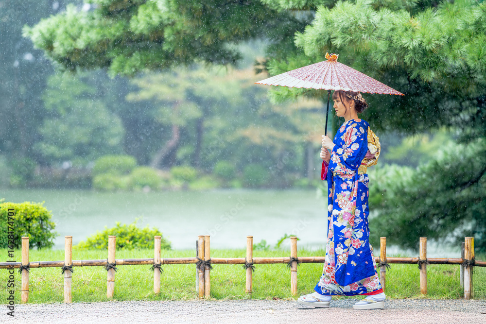 A local city in Japan on a rainy summer day. Japanese woman wearing a blue teenage kimono with a Japanese umbrella walking by a pond in a traditional garden.