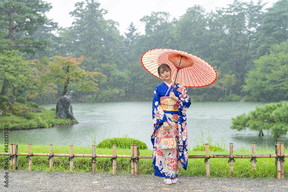 A local city in Japan on a rainy summer day. Japanese woman wearing a blue teenage kimono with a Japanese umbrella standing by a pond in a traditional garden.