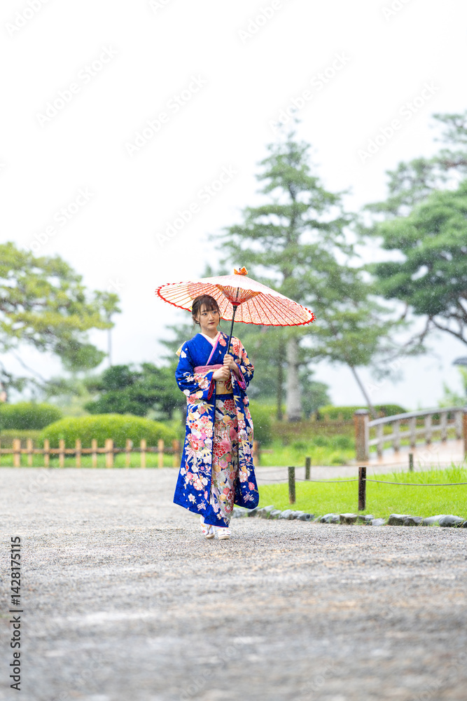 A local city in Japan on a rainy summer day. Japanese woman wearing a blue kimono for teenagers with a Japanese umbrella walking through a traditional garden.