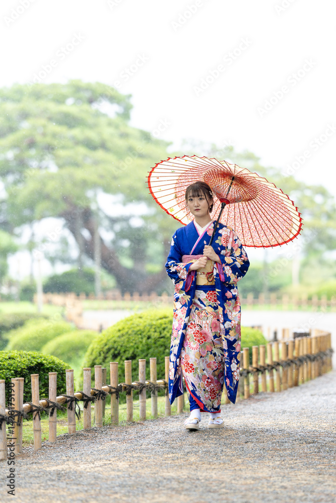A local city in Japan on a rainy summer day. Japanese woman wearing a blue kimono for teenagers with a Japanese umbrella walking through a traditional garden.