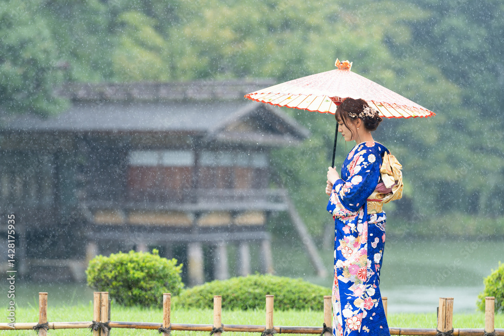 A local city in Japan on a rainy summer day. Japanese woman wearing a blue teenage kimono with a Japanese umbrella walking by a pond in a traditional garden.