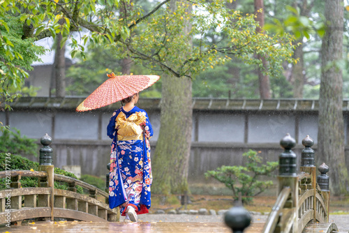 Obraz na płótnie A local city in Japan on a rainy summer day. Japanese woman wearing a blue teenage kimono with a Japanese umbrella walking along a traditional garden bridge. w salonie