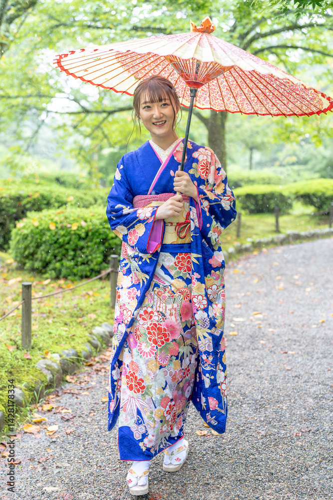 A local city in Japan on a rainy summer day. Japanese woman wearing a blue kimono for teenagers with a Japanese umbrella walking through a traditional garden.