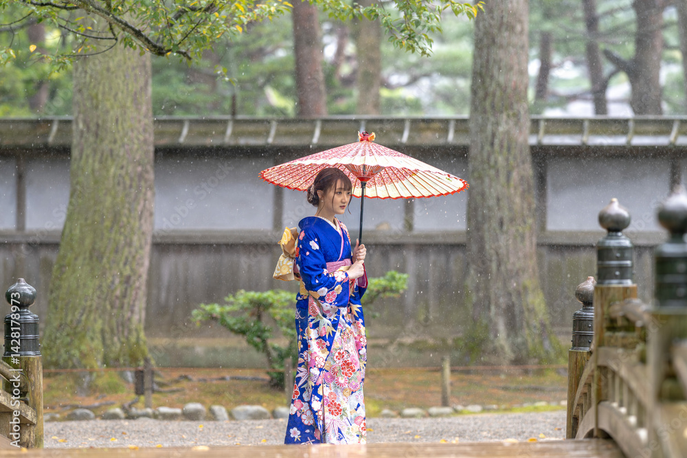 A local city in Japan on a rainy summer day. Japanese woman wearing a blue teenage kimono with a Japanese umbrella walking along a traditional garden bridge.