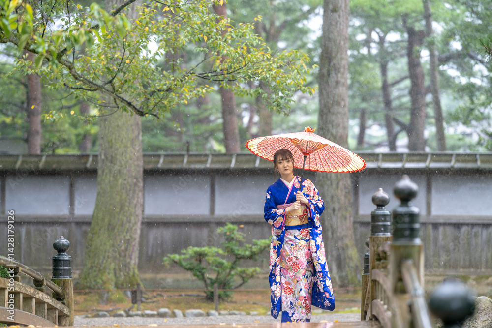 A local city in Japan on a rainy summer day. Japanese woman wearing a blue teenage kimono with a Japanese umbrella walking along a traditional garden bridge.