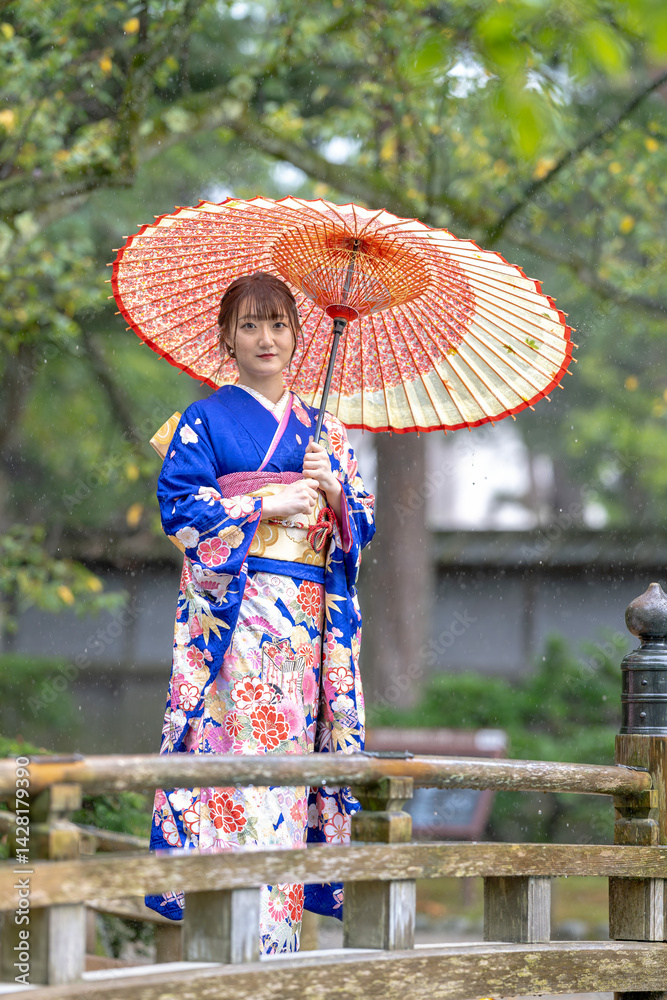 A local city in Japan on a rainy summer day. Japanese woman wearing a blue teenage kimono with a Japanese umbrella standing on a traditional garden bridge.