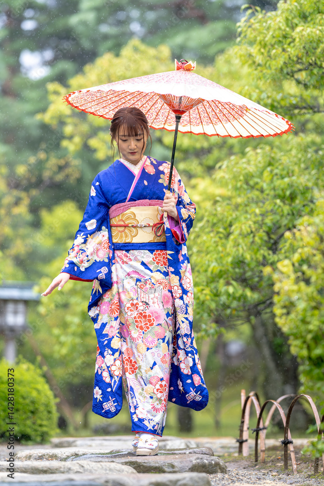 A local city in Japan on a rainy summer day. Japanese woman wearing a blue kimono for teenagers with a Japanese umbrella walking through a traditional garden.