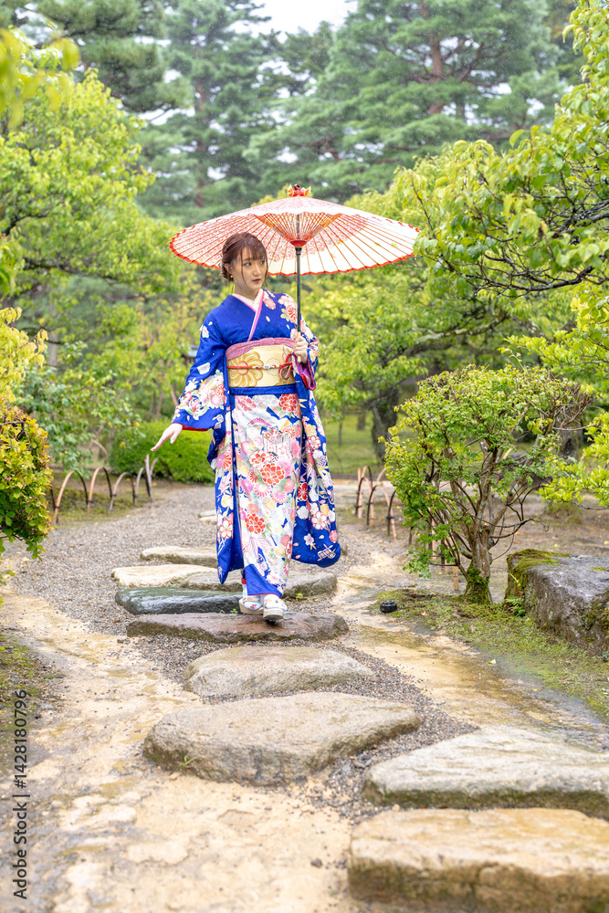 A local city in Japan on a rainy summer day. Japanese woman wearing a blue kimono for teenagers with a Japanese umbrella walking through a traditional garden.