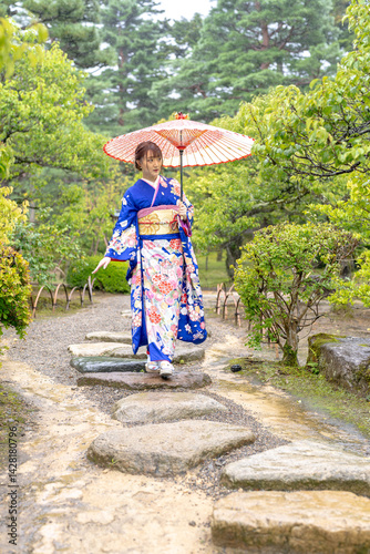Obraz na płótnie A local city in Japan on a rainy summer day. Japanese woman wearing a blue kimono for teenagers with a Japanese umbrella walking through a traditional garden. w salonie