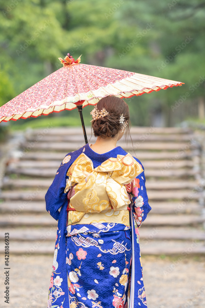 A local city in Japan on a rainy summer day. Japanese woman wearing a blue kimono for teenagers with a Japanese umbrella walking through a traditional garden. The back view.