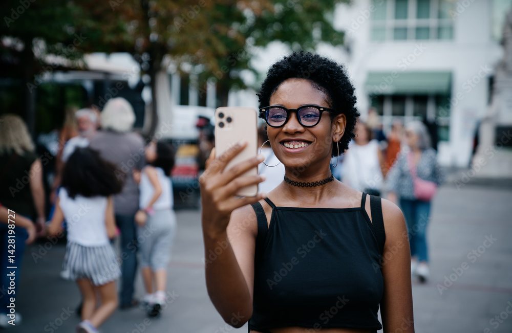 Happy young woman taking a selfie outdoors, standing confidently among people on a public street, smiling into her phone with natural charm and confidence