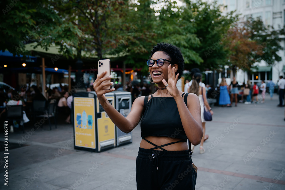 Smiling woman enthusiastically waving during video call on smartphone, surrounded by urban crowd and outdoor cafes in warm evening light