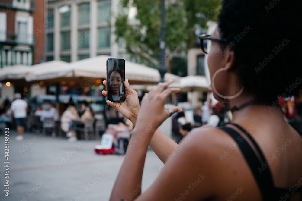 Young Black woman talking on video call, gesturing with hand while holding smartphone, enjoying online conversation at vibrant city terrace surrounded by locals and tourists