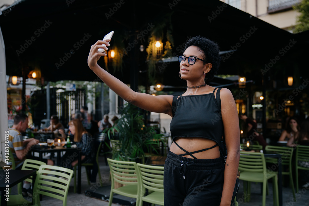 Young stylish Black woman takes a selfie outside cozy evening café, capturing content for vlog or social media, surrounded by people dining in ambient urban atmosphere