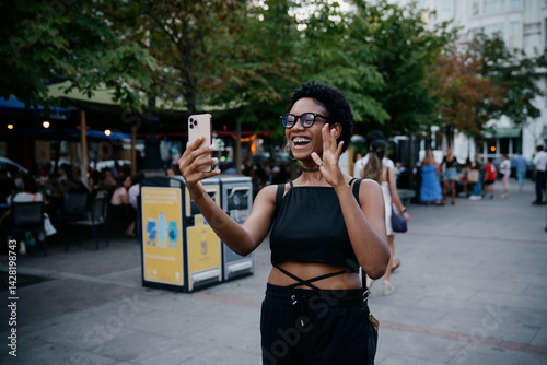 Obraz na płótnie Smiling woman enthusiastically waving during video call on smartphone, surrounded by urban crowd and outdoor cafes in warm evening light w salonie