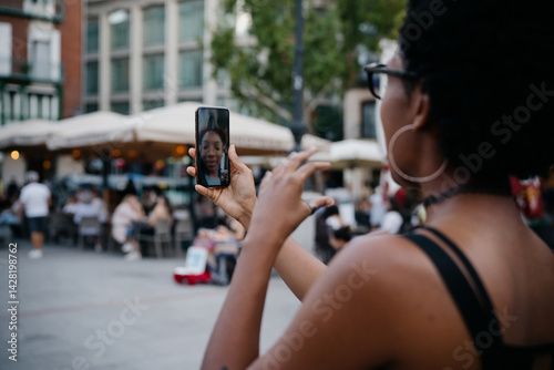 Obraz na płótnie Young Black woman talking on video call, gesturing with hand while holding smartphone, enjoying online conversation at vibrant city terrace surrounded by locals and tourists w salonie