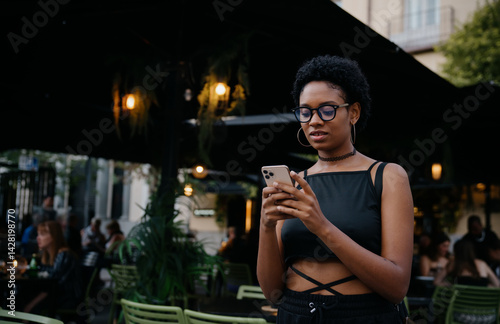 Obraz na płótnie Trendy woman holding smartphone while checking messages in outdoor evening café, blending technology with relaxed ambiance and modern lifestyle in urban nightlife scene w salonie