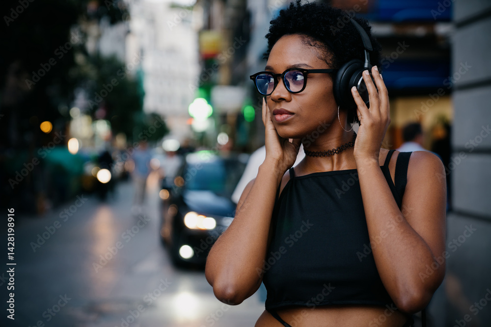Focused young woman listens to streaming audio with wireless headphones, standing on lively urban street, immersed in music, podcast, or audiobook while enjoying the city vibe